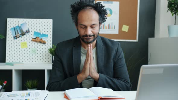 Pensive Middle Eastern Man Holding Hands in Namaste Gesture Sitting at Desk with Eyes Closed
