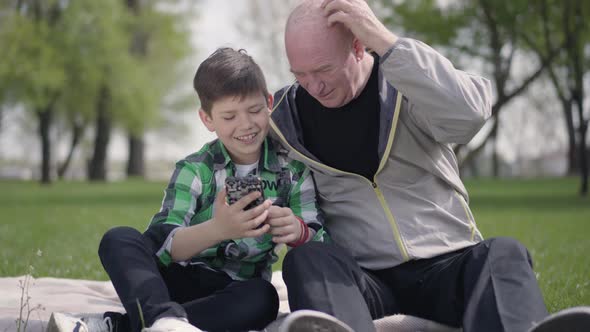 Senior Man Sitting with His Grandson on the Blanket in the Park, Looking in Cellphone