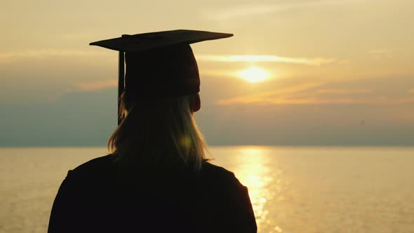 A Woman in a Graduate Robe and in a Cap Looks at the Sunrise Over the Sea