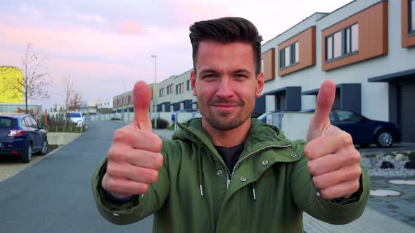 A Man Stands Outside, Shows Thumbs Up and Smiles at the Camera, Houses in the Background