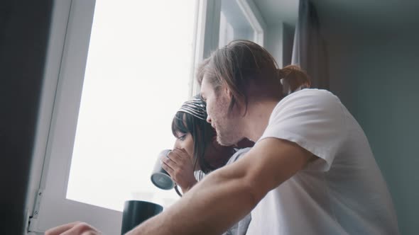 Young Happy Multiracial Couple Drinking Coffee or Tea and Looking Through the Window