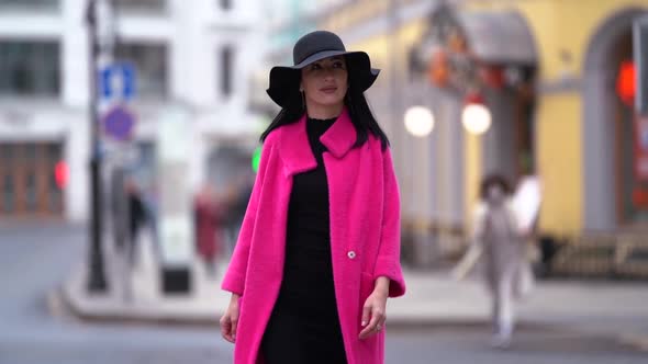 A Stylish Black-haired Woman in a Pink Coat and Black Hat Walks and Poses on a Blurry City Street