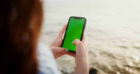 A Woman is Typing on Smartphone with a Green Screen Sitting Seashore