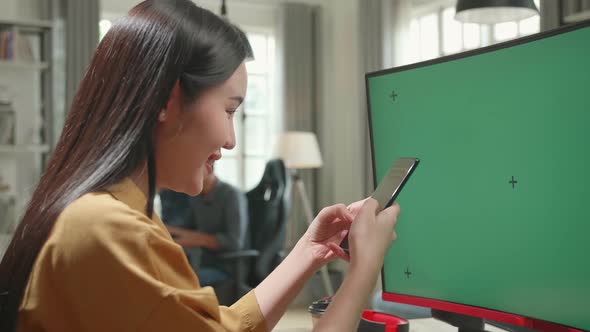 Asian Female Works And Using Mobile Phone With Mock Up Green Screen Computer Display In Office