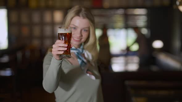 Happy Young Woman Toasting Stretching Beer Glass to Camera Smiling Standing in Pub Indoors