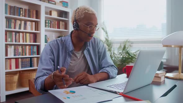 Serious Darkskinned Man is Talking on Headset and Making Notes in Documents