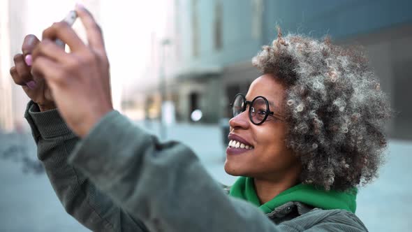 Smiling adult African woman wearing casual clothes taking photo of building on phone