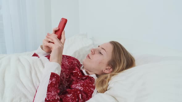 The Girl Lies with Her Phone in Her Hands in a Comfortable Cozy Fresh Bed in Her Red Pajamas