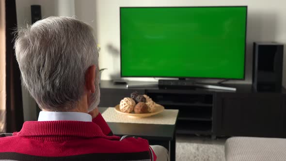 An Elderly Man Sits on a Sofa in Living Room and Watches TV with a Green Screen - Focus on the Head