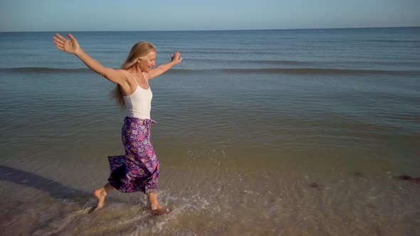 With arms outstretched, a slow motion clip of a mature woman splashing through the water on a beach