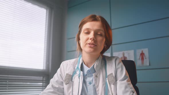 Young woman brunette doctor waves hand greeting patient at online meeting in local hospital room