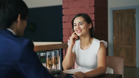 Cheerful Young Lady in Fancy Dress Is Speaking with Her Boyfriend on Date Sitting at Table Together