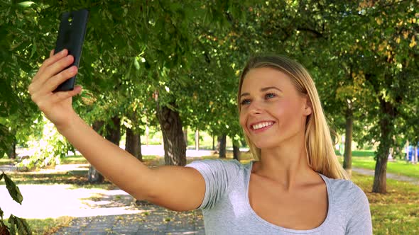 Young Pretty Blond Woman Photographs with Smartphone (Selfie) - Park with Trees in Background