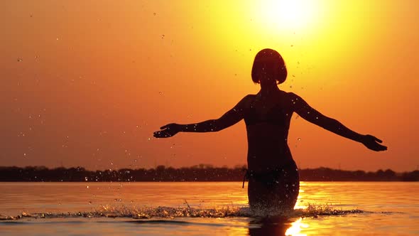 Silhouette of Woman at Sunset Raises Hands Up and Creating Splashes of Water. Slow Motion