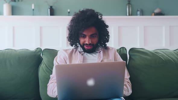 Young Overjoyed Arabian Man with Laptop Waves Hands Sits on Sofa in Living Room