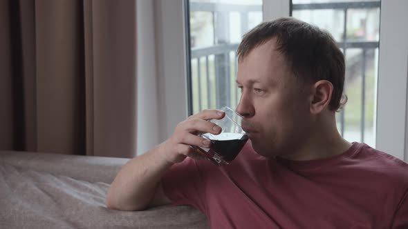 Man with a Serious Expression is Drinking From a Glass Sitting on the Sofa Window Background