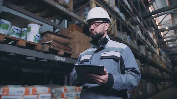 Warehouse Worker in a White Helmet Walks Along the Rows with Boxes