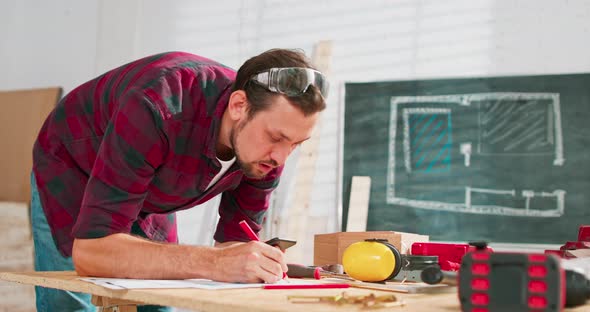 A Handsome Carpenter in a Flannel Shirt Counts the Dimensions on a Smartphone in a Carpentry