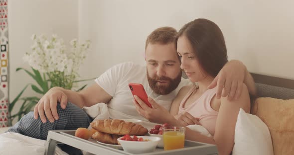 Young Man and Woman Looking at Phone Screen and Having Good News While Lying on Bed. Happy Couple