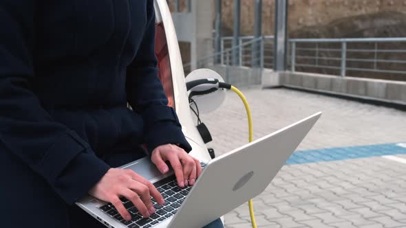 A Woman Sits on the Trunk of an Electric Car with a Laptop and Waits for the Car to Charge in the