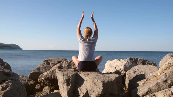 Young Woman Sits on the Stones By the Sea and Practices Yoga in the Early Morning