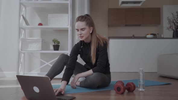 Girl Watching Training Videos with Sports Training at Home. Sitting on a Mat Near a Dumbbell