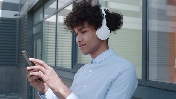 Ethnic Man in Headphones Looking at Smartphone Near Building in Town
