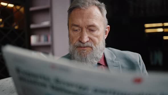 Portrait of Concentrated Mature Bearded Man Wearing Suit Reading Newspaper Sitting at Luxury Dark