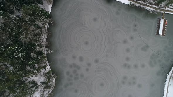 Aerial shot of frozen lake with small lake house surrounded by forest at winter