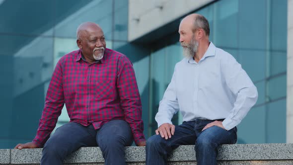 Two Diverse Bearded Elderly Male Friends in Casual Clothes Shirts and Jeans Sitting Outdoors