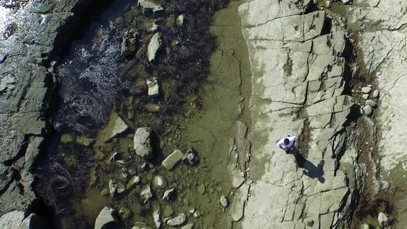 Aerial birds eye view shot of a young man running on a rocky ocean beach shoreline.