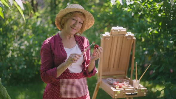 Portrait of Cheerful Senior Woman Posing with Painting Brush and Strawberry at Easel Outdoors