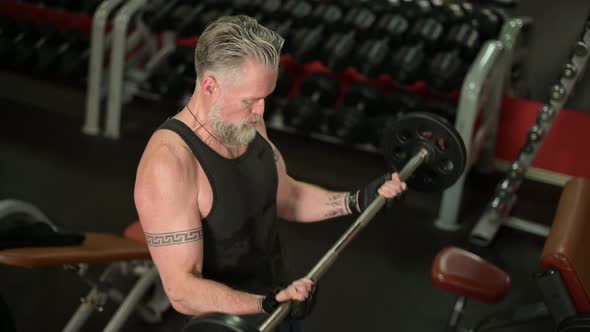 A gray-haired, solid tattooed man lifts a heavy barbell to his biceps
