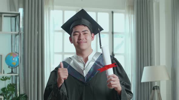 Excited Asian Man Holding A University Certificate Smiling And Thumb Up To Camera On The Living Room