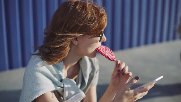 Beautiful Young Woman Uses a Smartphone on the Street and Licks Colored Lollipop