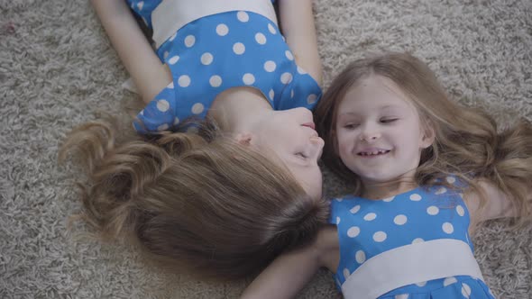 Top View of Two Pretty Caucasian Twin Sisters Lying on Soft Carpet and Smiling. Camera Moving Around