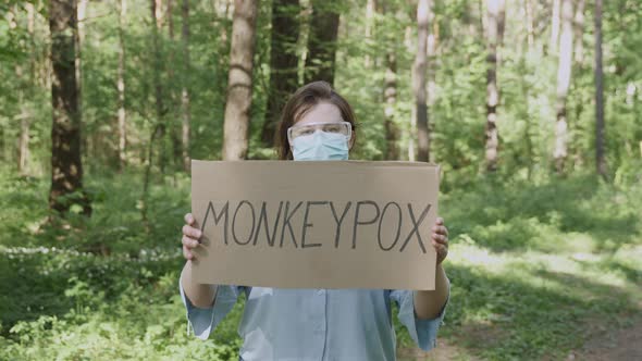 Woman in Mask Glasses Stands with Sign Monkeypox in Green Sunny Forest