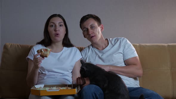 Lady Sits By Man on Sofa Eats Pizza Feeding Husband and Cat