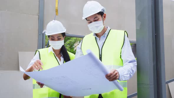 Asian construction workers people wearing protective face mask onsite and holding blueprint drawing.