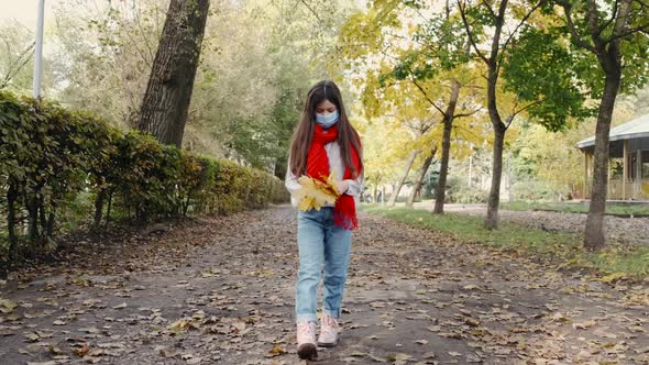 Stylish Girl in Mask and Red Scarf Is Picking Up Leaves While Walking