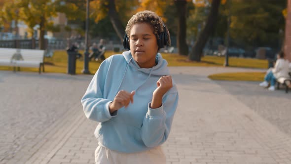 Young Smiling African Woman Listening Music with Headphones and Dancing Walking Down City Street