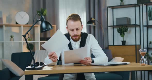 Bearded Man Sitting at His Workplace at Home and Revisioning Different Reports