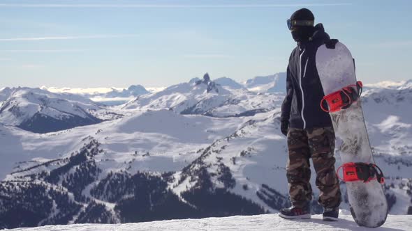 A young man snowboarder standing with his snowboard on a snow covered mountain.