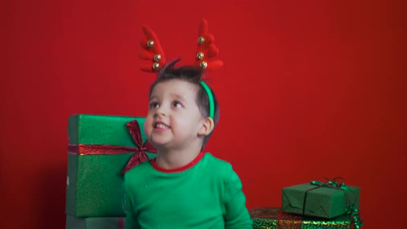 Child in a Green Dwarf Costume Sits on Gift Boxes with Deer Antlers