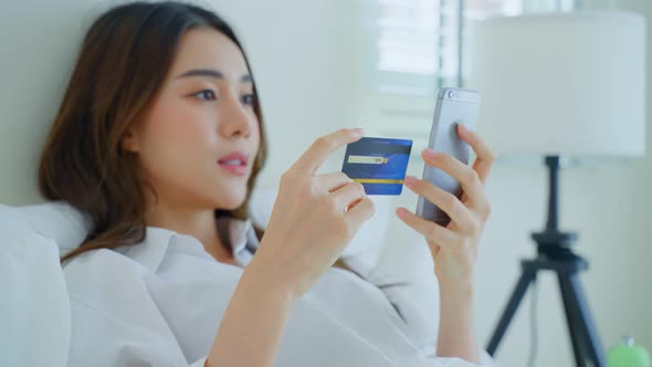 Asian attractive woman enjoy shopping online on bed at home in morning holding credit card.