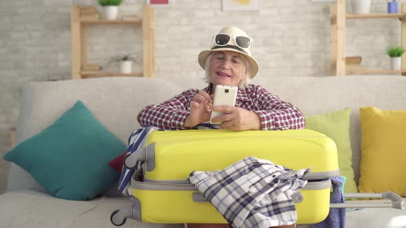 Smiling Old Woman in a Hatuses a Smartphone with a Suitcase for Travel in His Hands Sitting on the