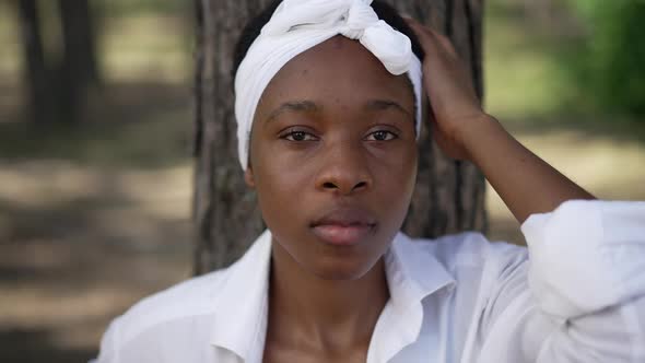 Closeup Front View of Confident Young African American Woman Looking at Camera Leaning on Tree Trunk