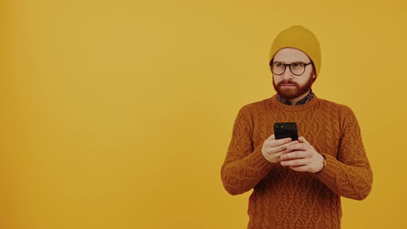 European Hipster in a Beanie and Orange Sweater Addicted to Using His Black Smartphone Studio Shot