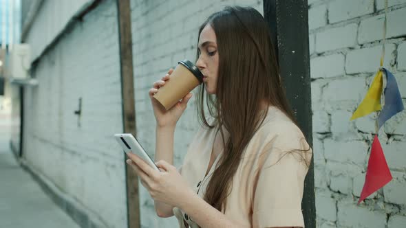 Pensive Young Woman Touching Smartphone Screen and Drinking to Go Coffee Outdoors in City Street