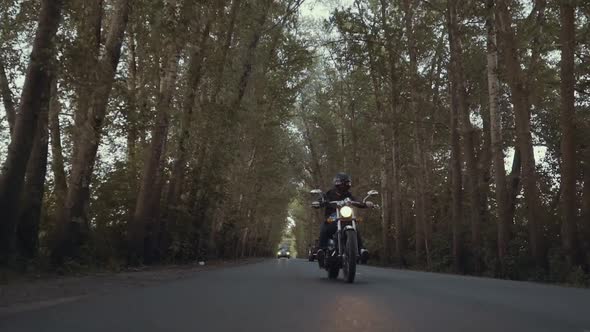 A Guy in a Black Leather Jacket and Helmet Riding a Classic Motorcycle on a Forest Road. Close-up on